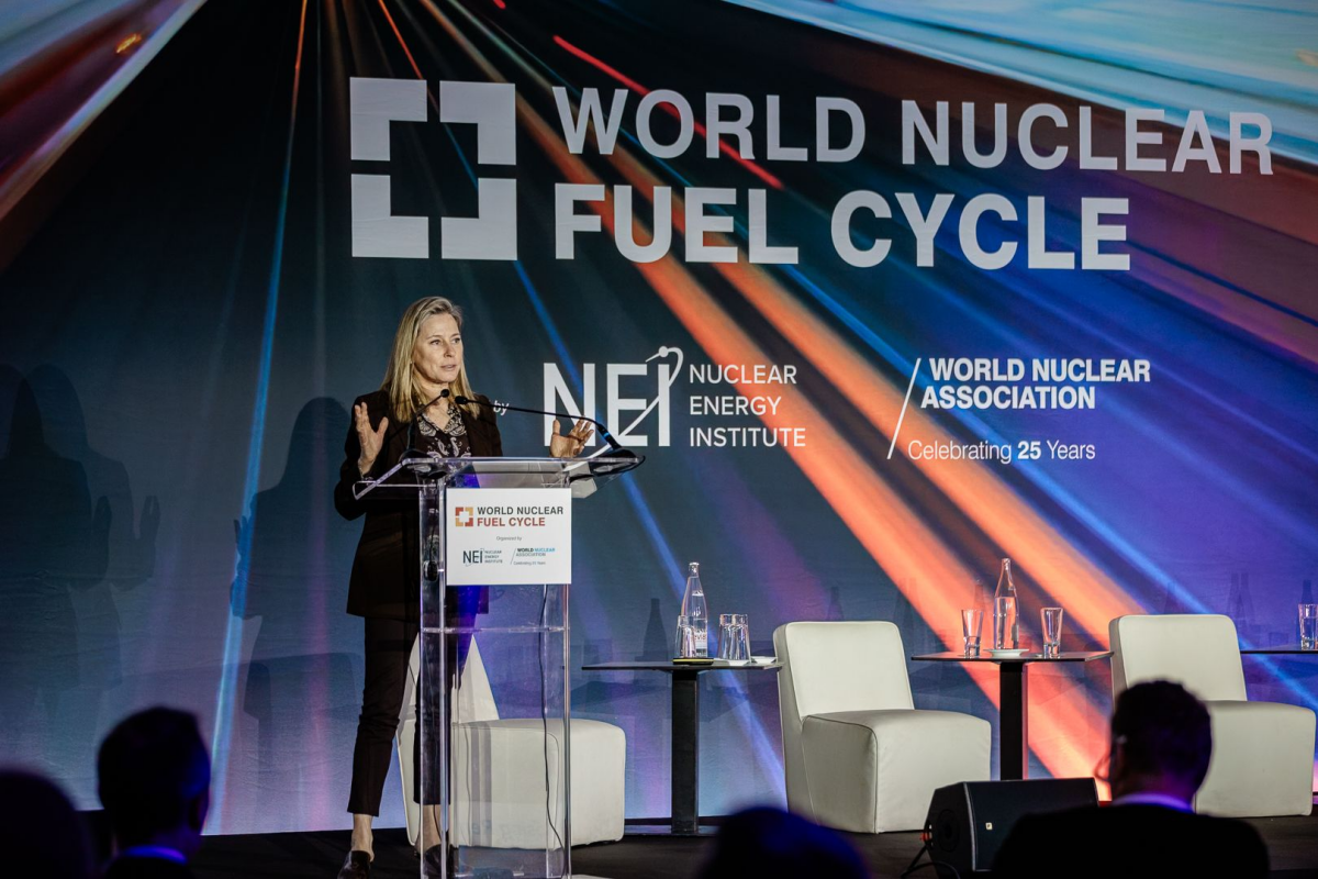 Female speaker at a clear podium on a stage with a large World Nuclear Fuel Cycle backdrop and logos (NEI, World Nuclear Association) in colorful lighting.
