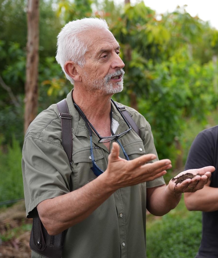 Older man with white hair and beard holding soil in cupped hands while speaking outdoors in a garden setting.