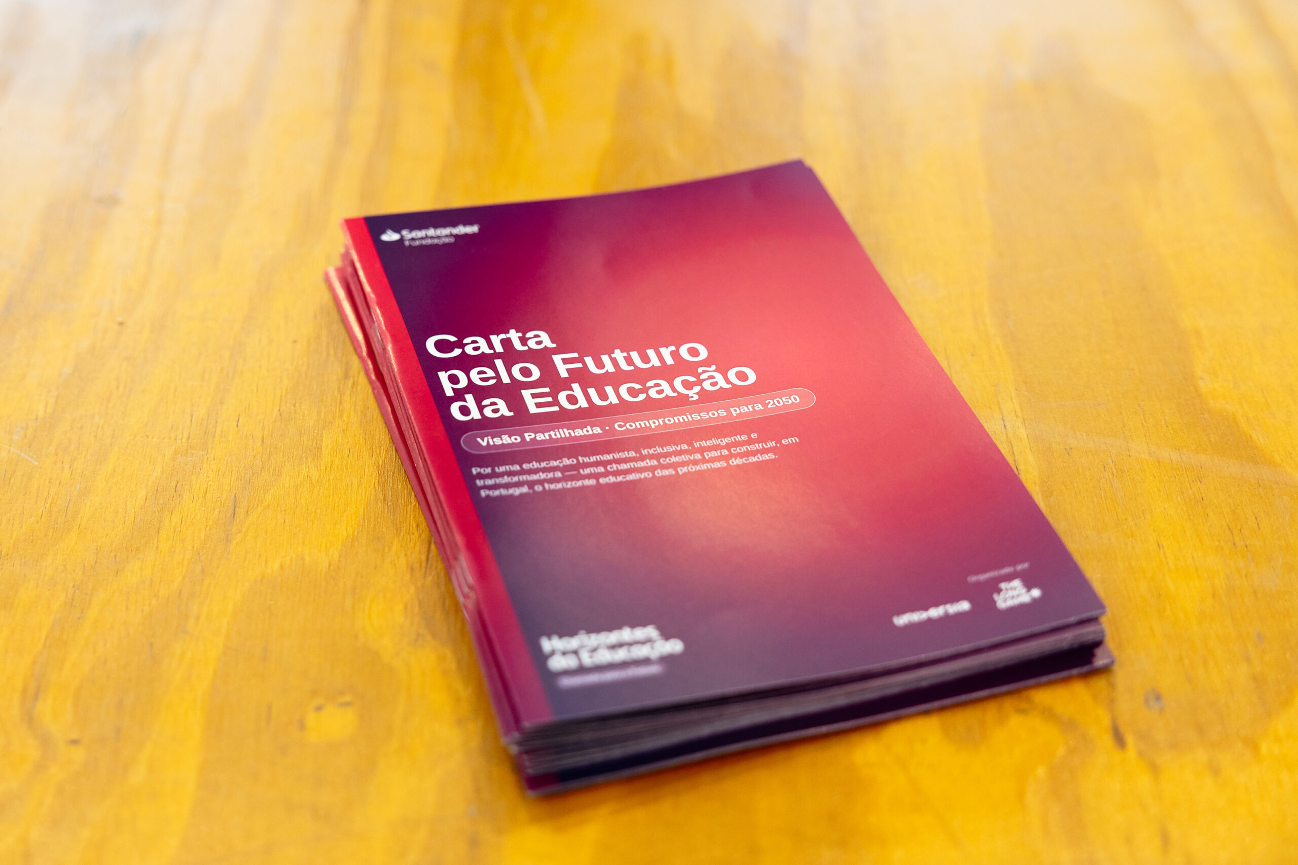 Red gradient booklet on a wooden table, titled 'Carta pelo Futuro da Educação' with white text.