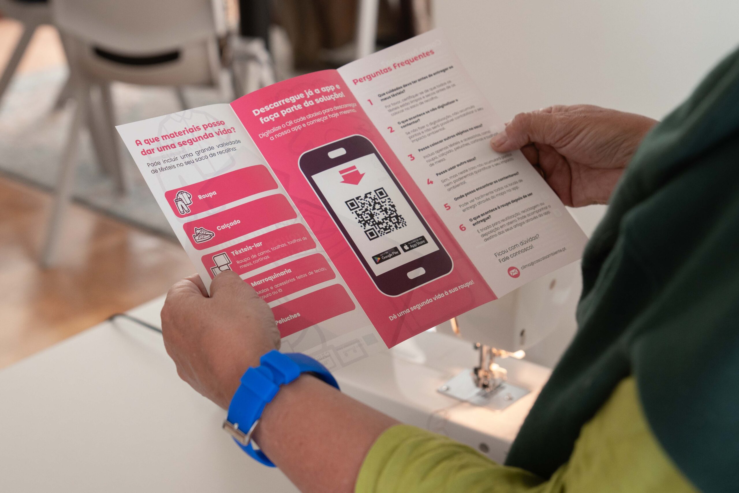 Person holding a pink brochure with a smartphone graphic and QR code, read by a seated person wearing a blue watch.