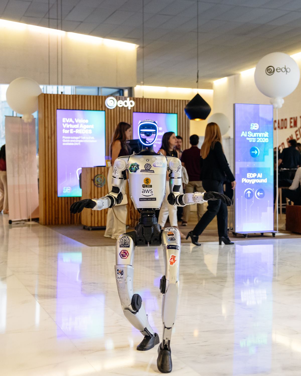 Humanoid robot standing in a lobby at a tech expo, with attendees and booth displays in the background.