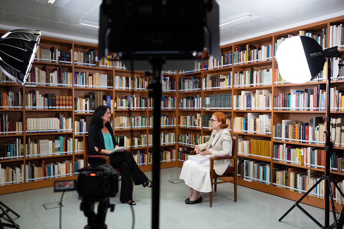 Two women in a library interview setup, facing each other with notebooks; film lights and cameras are visible in the scene.