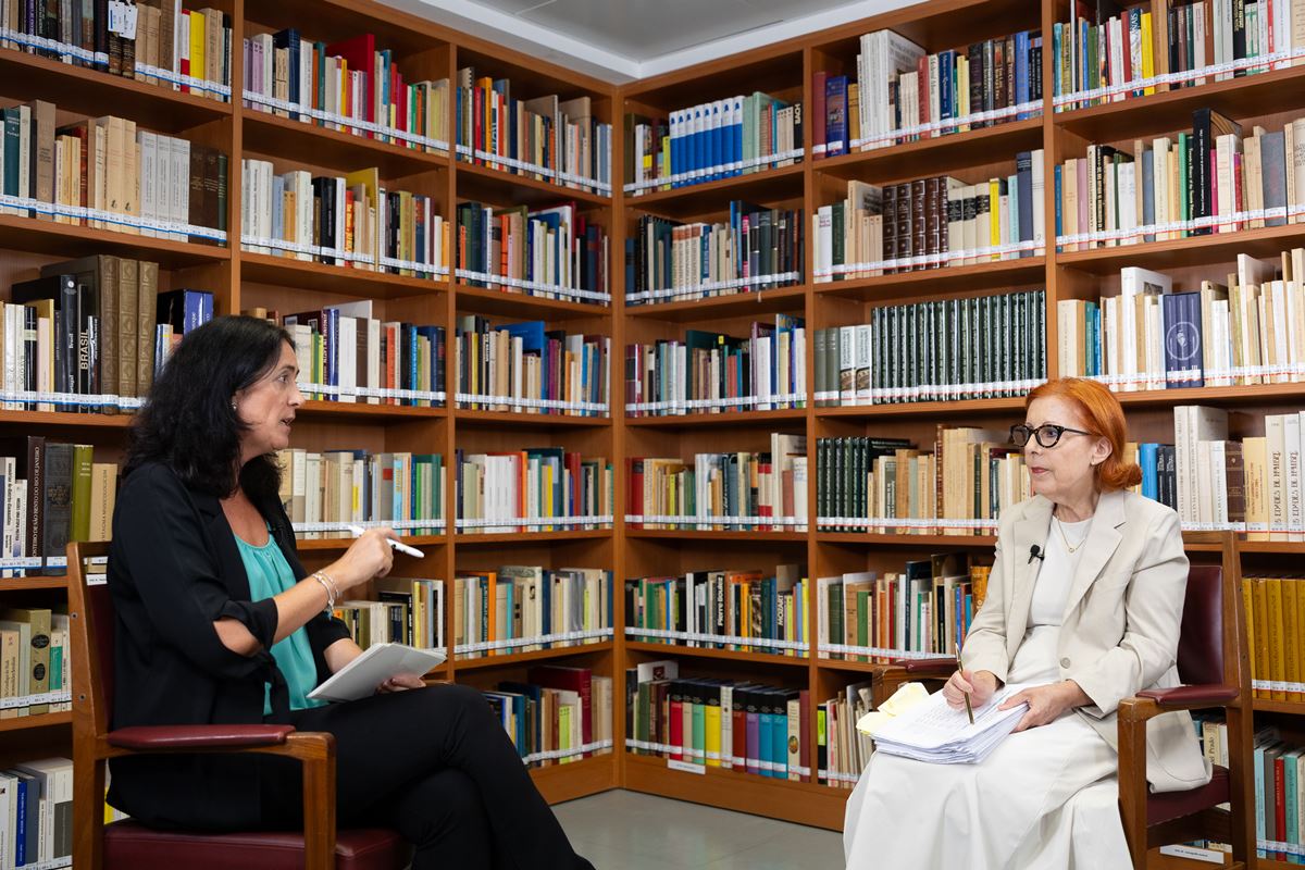 Two women sit in a library, one interviewee holding papers while the other speaks with a notebook in hand.